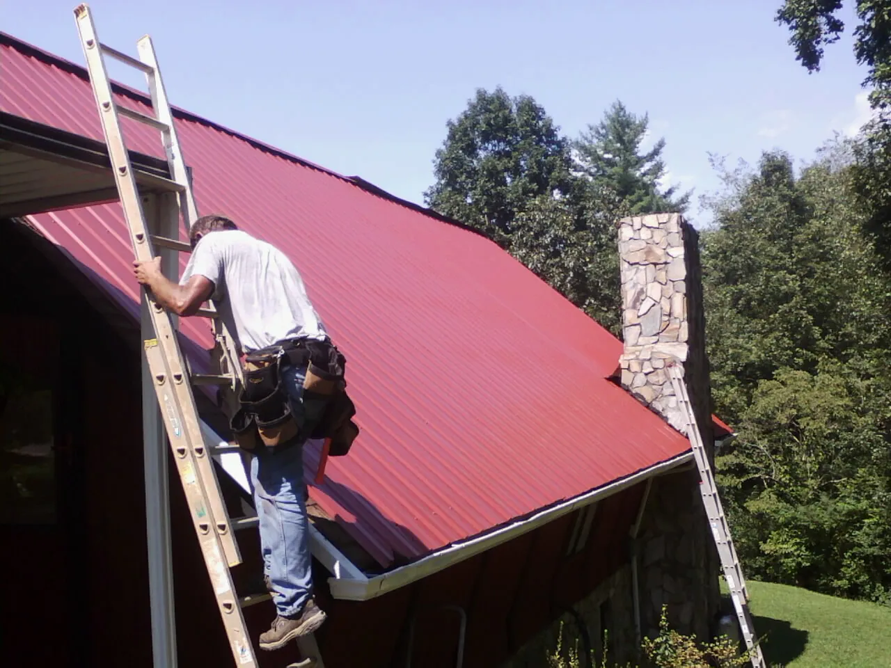 Roofer on a ladder beside a red metal roof and stone chimney — Soffit & Fascia Repair by Roofing Brothers in Klamath Falls